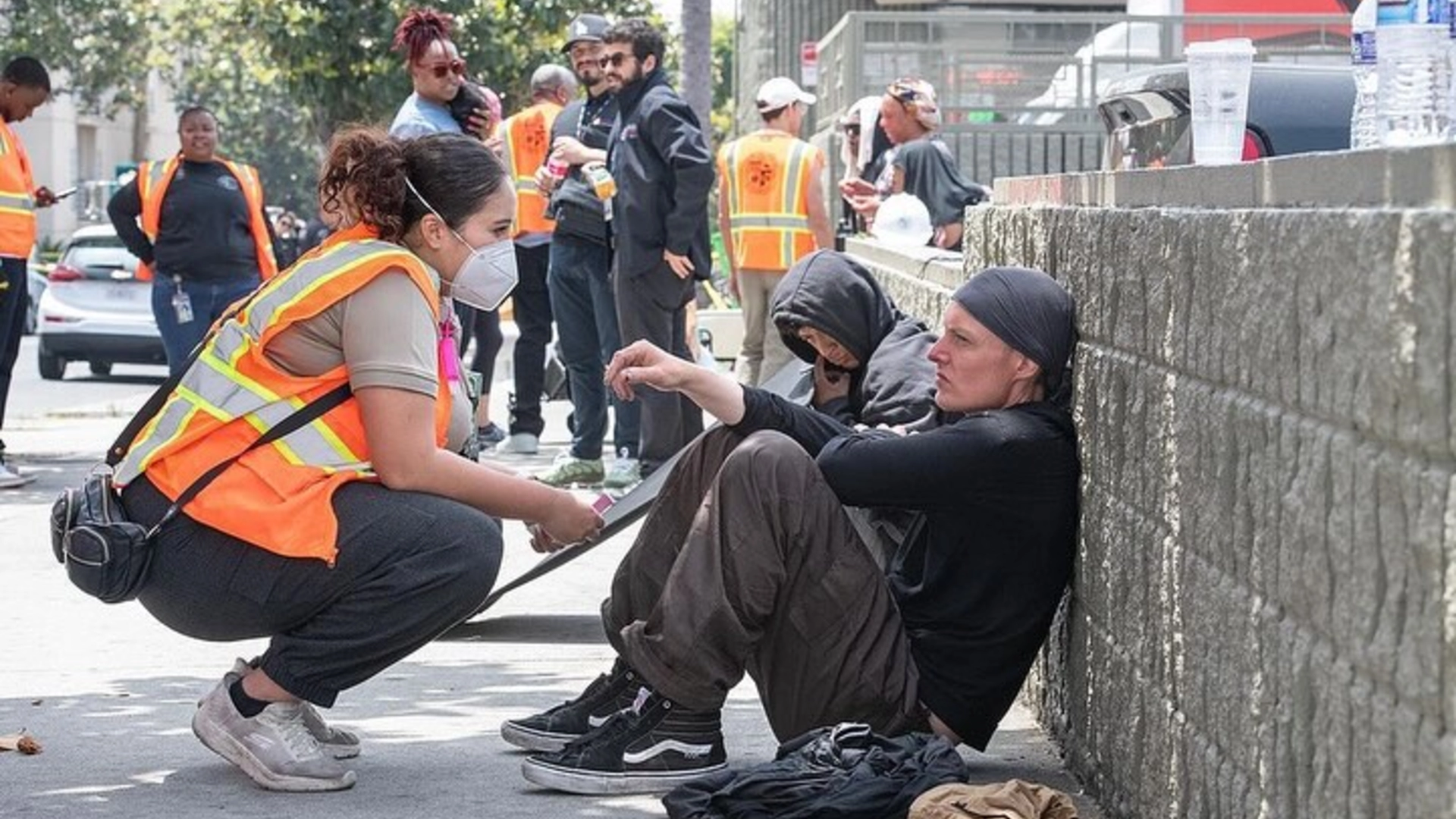 Woman wearing orange vest looking at two people leaning against wall